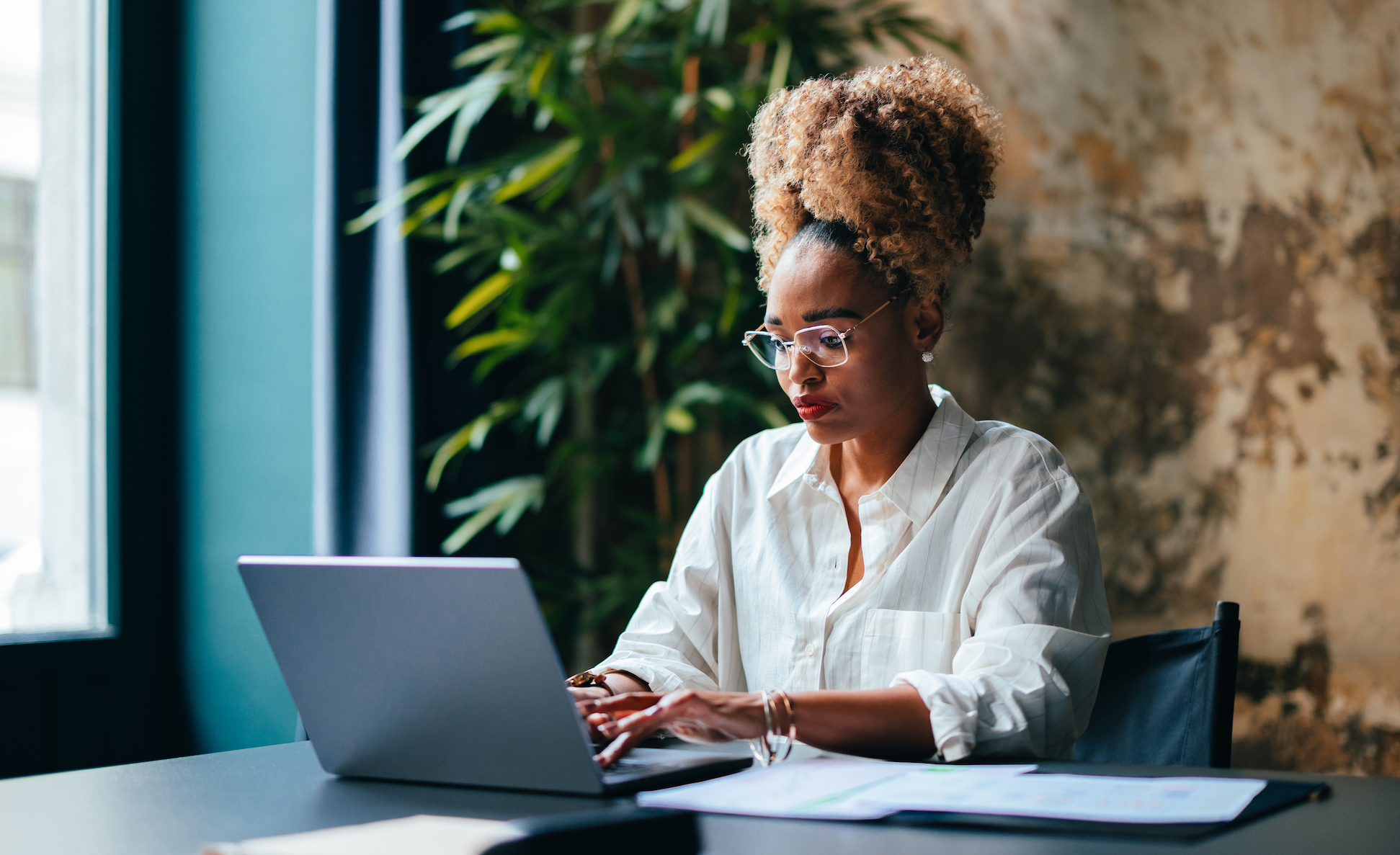 A young black female entrepreneur uses a laptop at a desk with papers spread out